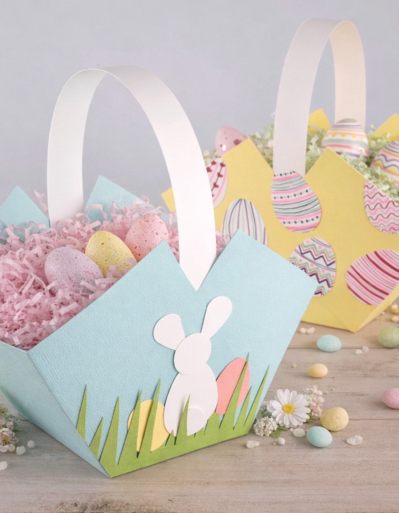 Two handmade paper Easter baskets filled with shredded paper and eggs, one with bunny and grass decoration and one with colorful egg prints.