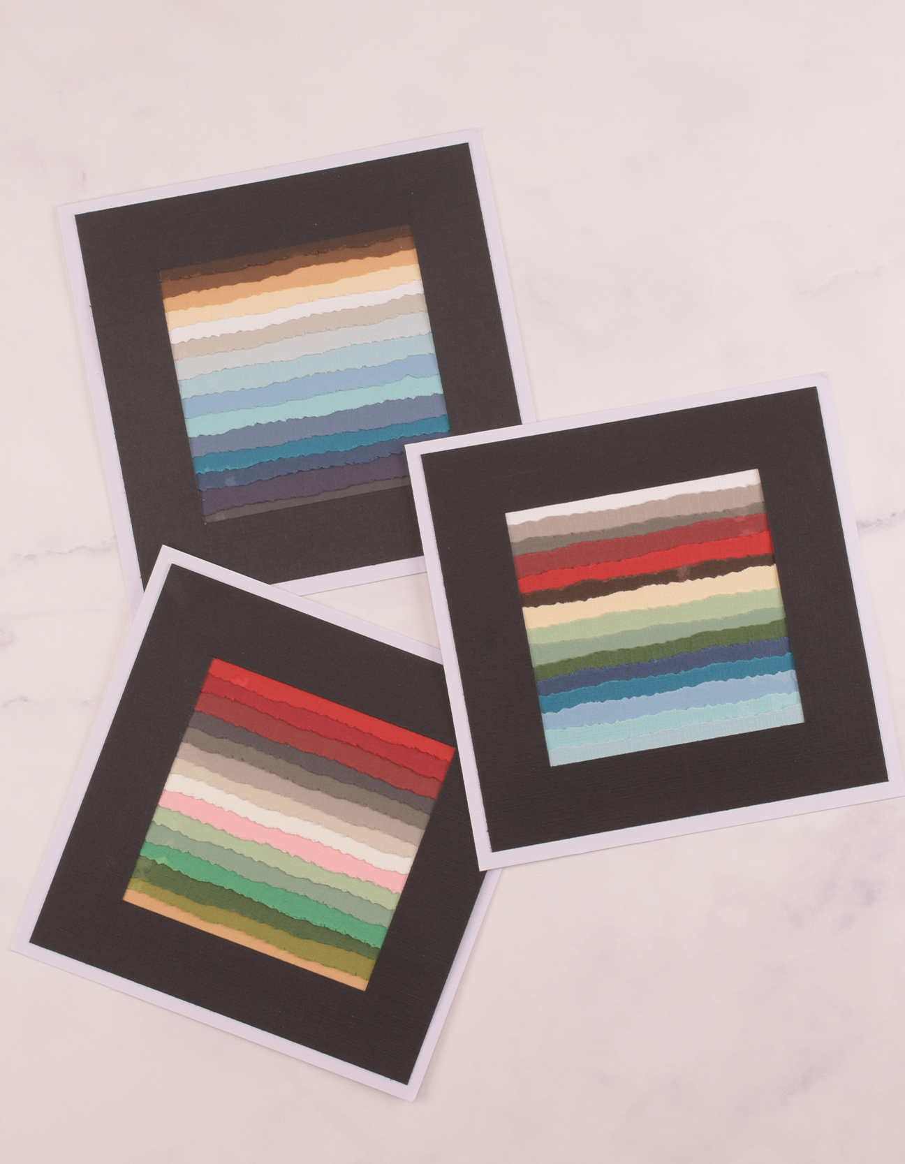 Drei fertige DIY-Schichtbilder mit gerissenen farbigen Papierstreifen im schwarzen Passepartout auf weißem Karton.