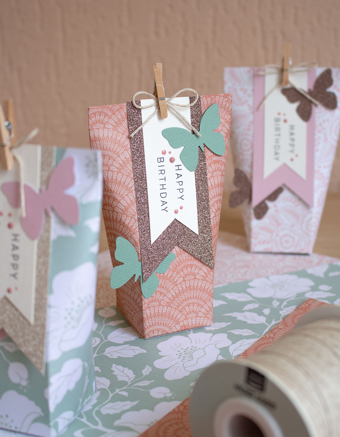 Drei wunderschön dekorierte DIY-Geschenkboxen mit rosa Blumendruckpapier, Schnur-Schleifen und Glitzer-Schmetterlingen, bereit zum Verschenken