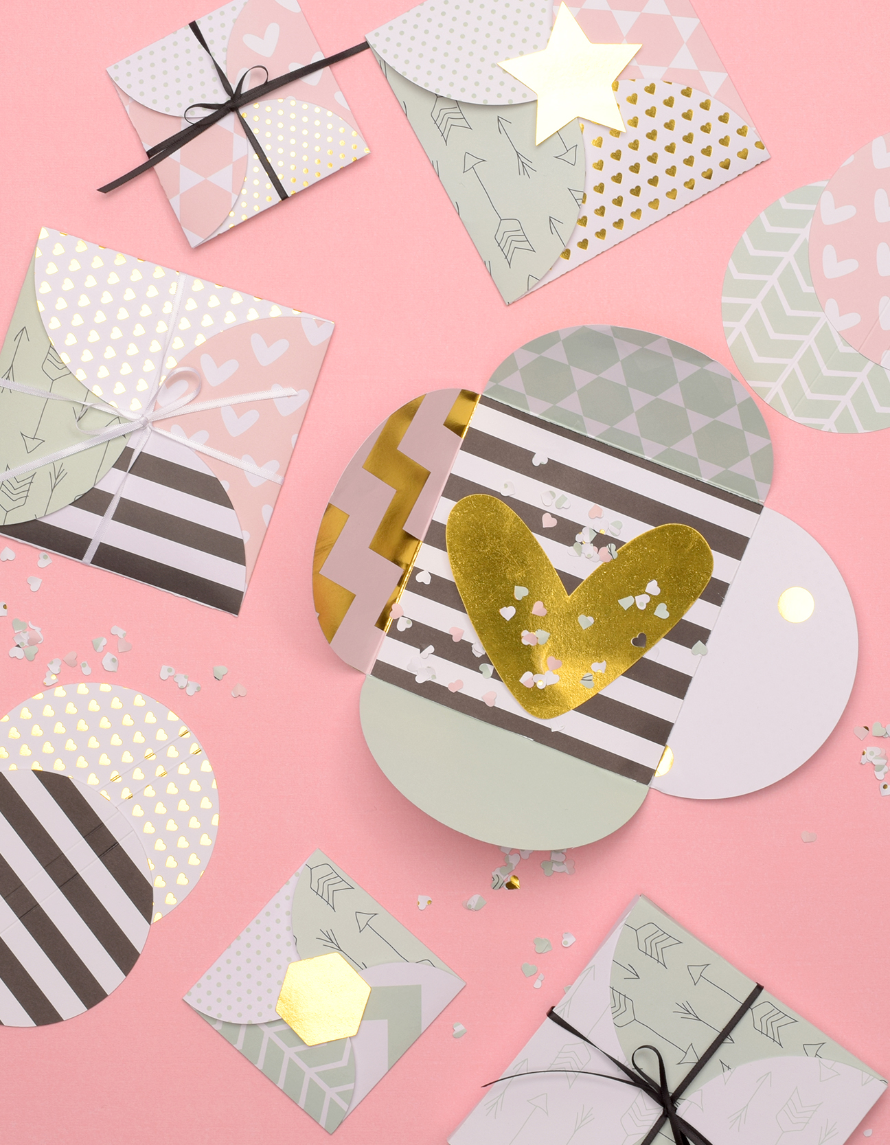 Afgewerkte handgemaakte gevouwen cadeauenvelopjes en mini cadeaudoosjes van papier met confetti, lintjes en pastel dessinpapier op een roze achtergrond.