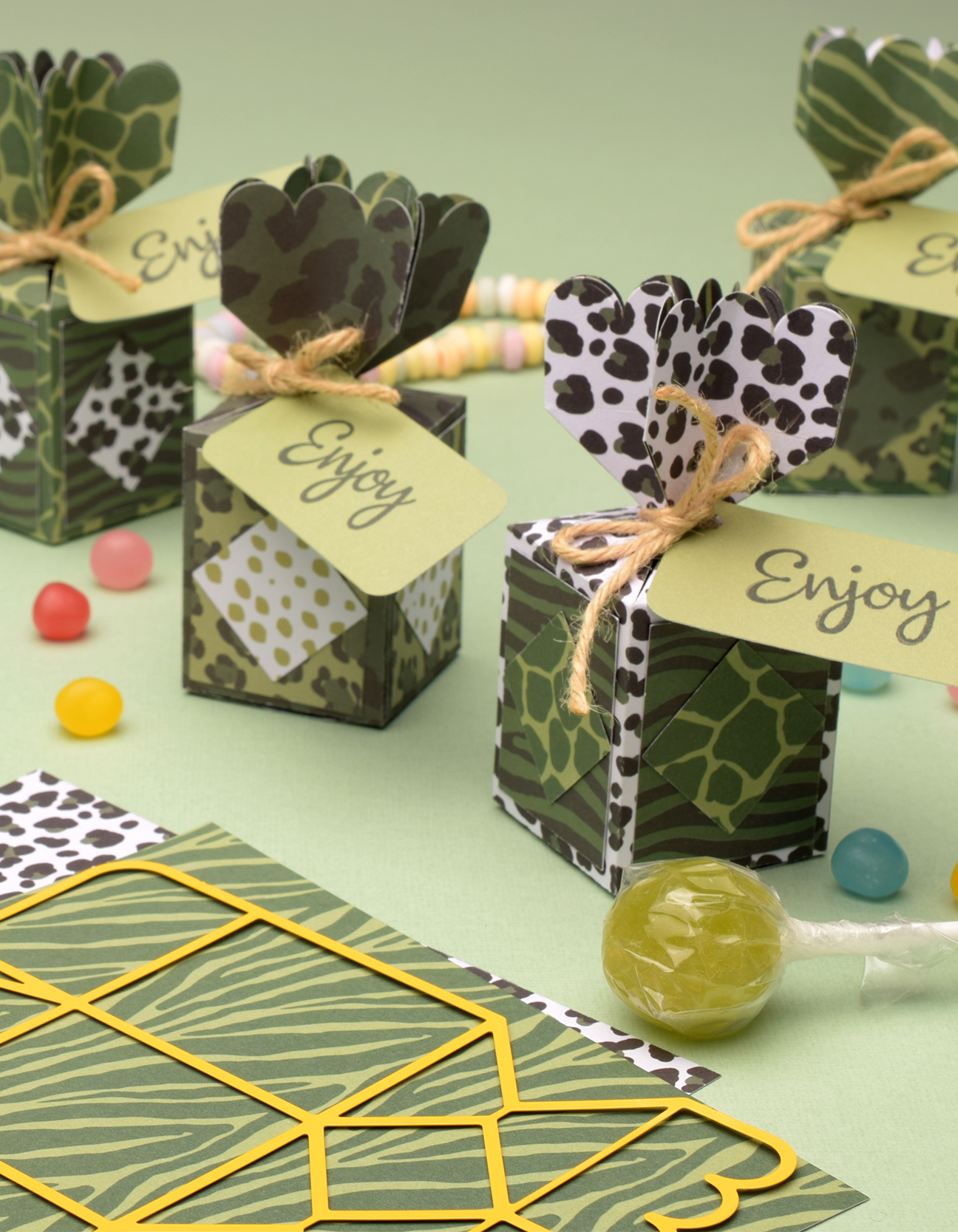 Cajas de regalo safari hechas a mano con estampados animales, lazos de cuerda y etiquetas “Enjoy”, presentadas con dulces y un troquel sobre fondo verde suave.