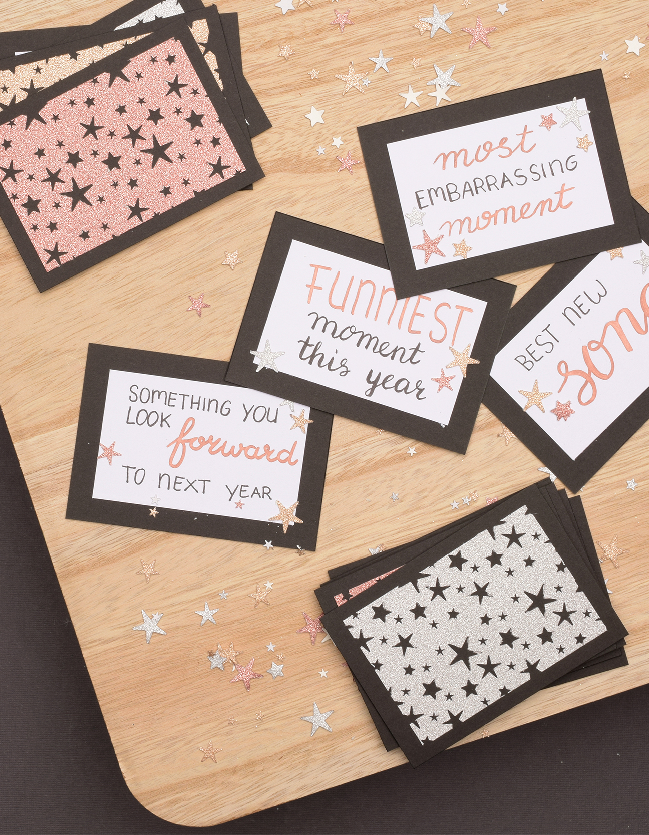 Biglietti di riflessione di Capodanno fatti a mano con domande in handlettering, bordi neri e decorazioni di stelle glitter, disposti su un vassoio di legno.