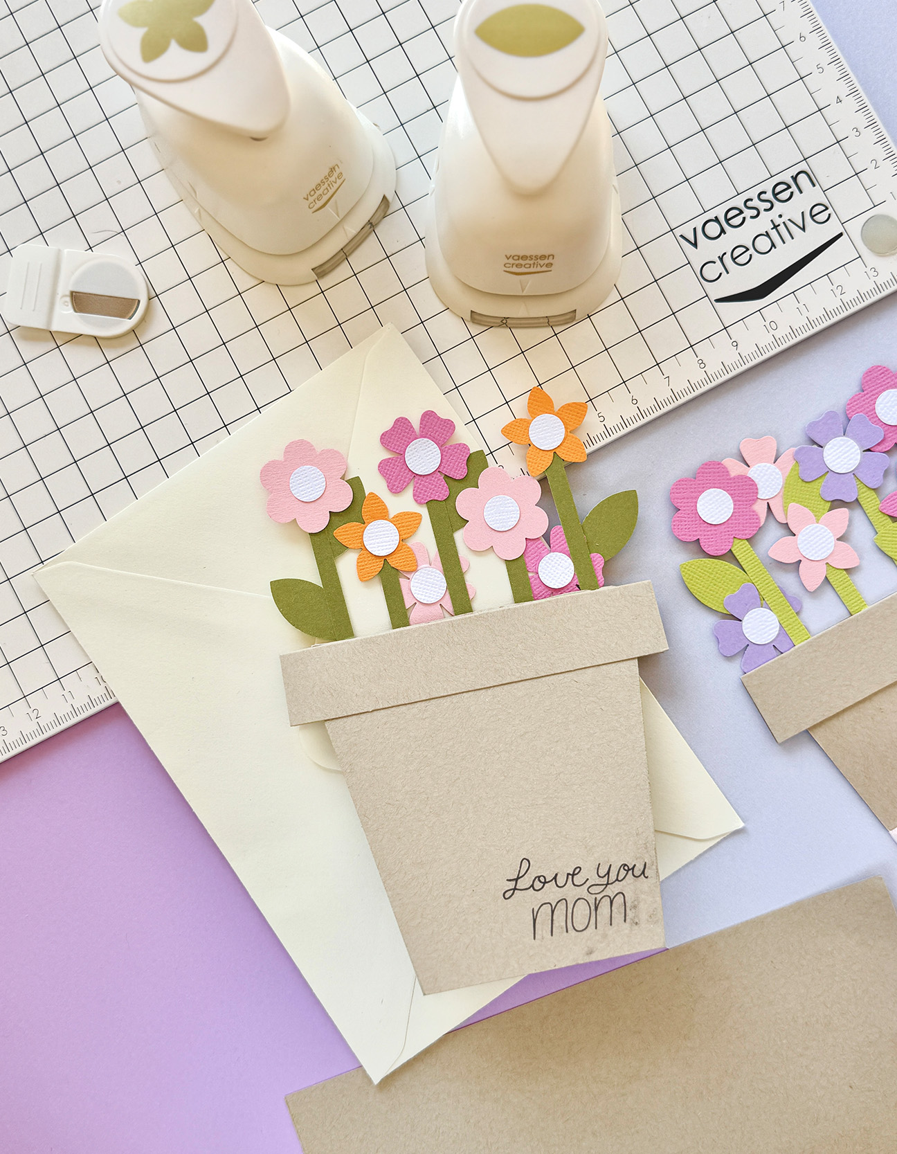 Biglietto artigianale per la Festa della Mamma a forma di vaso di fiori, con fiori di carta colorati, busta e fustelle, presentato come un allegro regalo fai da te per la mamma.