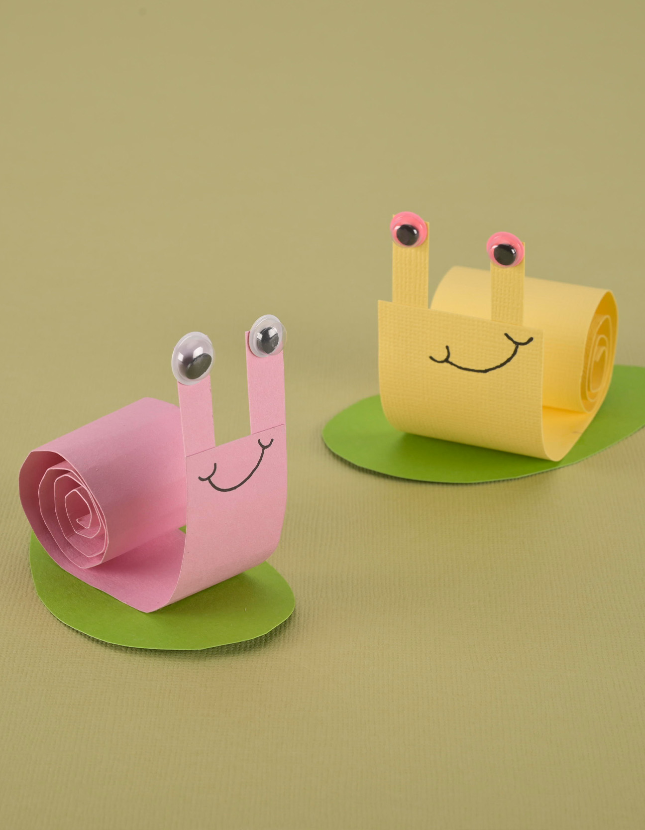 Deux escargots colorés en papier réalisés avec du cardstock enroulé et des yeux mobiles sur des feuilles vertes pour une activité manuelle de printemps amusante.