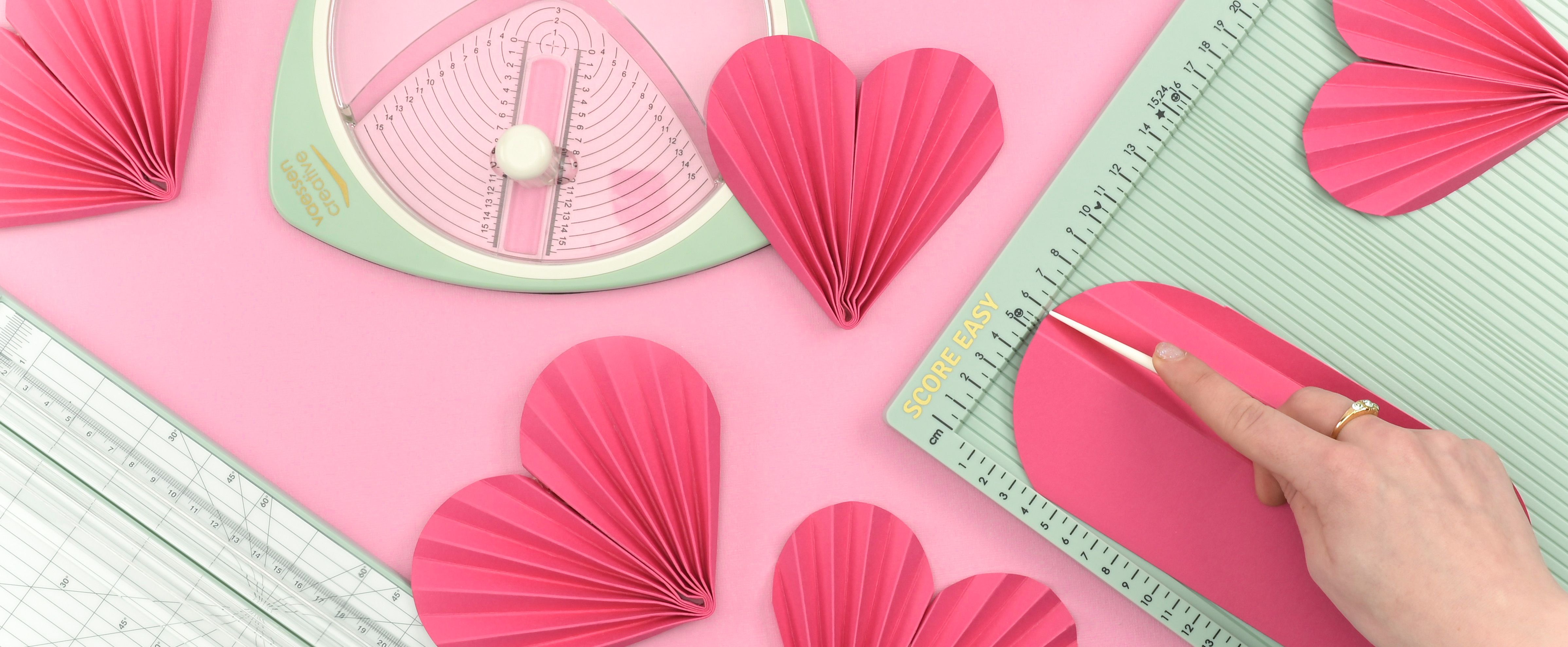 DIY paper hearts being scored and folded on a scoring board, with pink folded heart shapes arranged around the workspace.