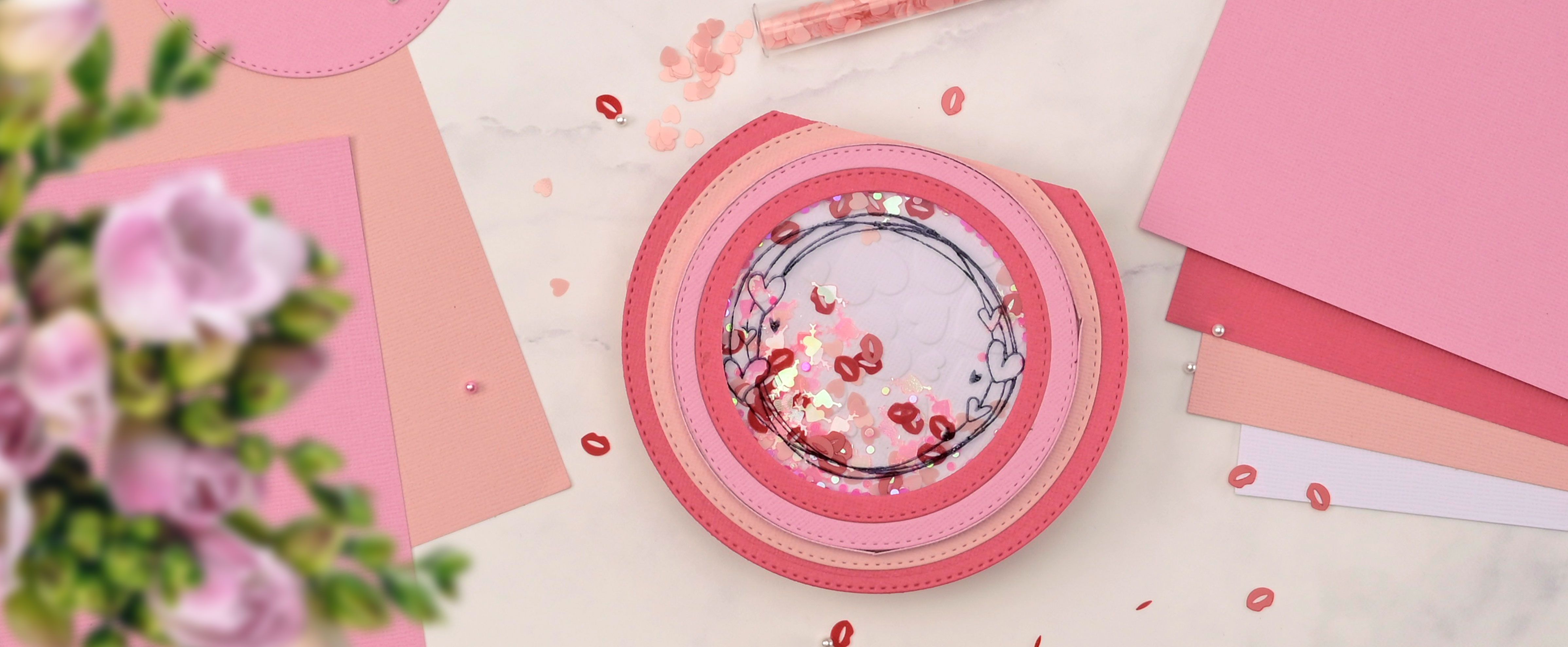 Handmade round shaker card in pink shades with heart confetti, surrounded by pink paper and craft supplies.