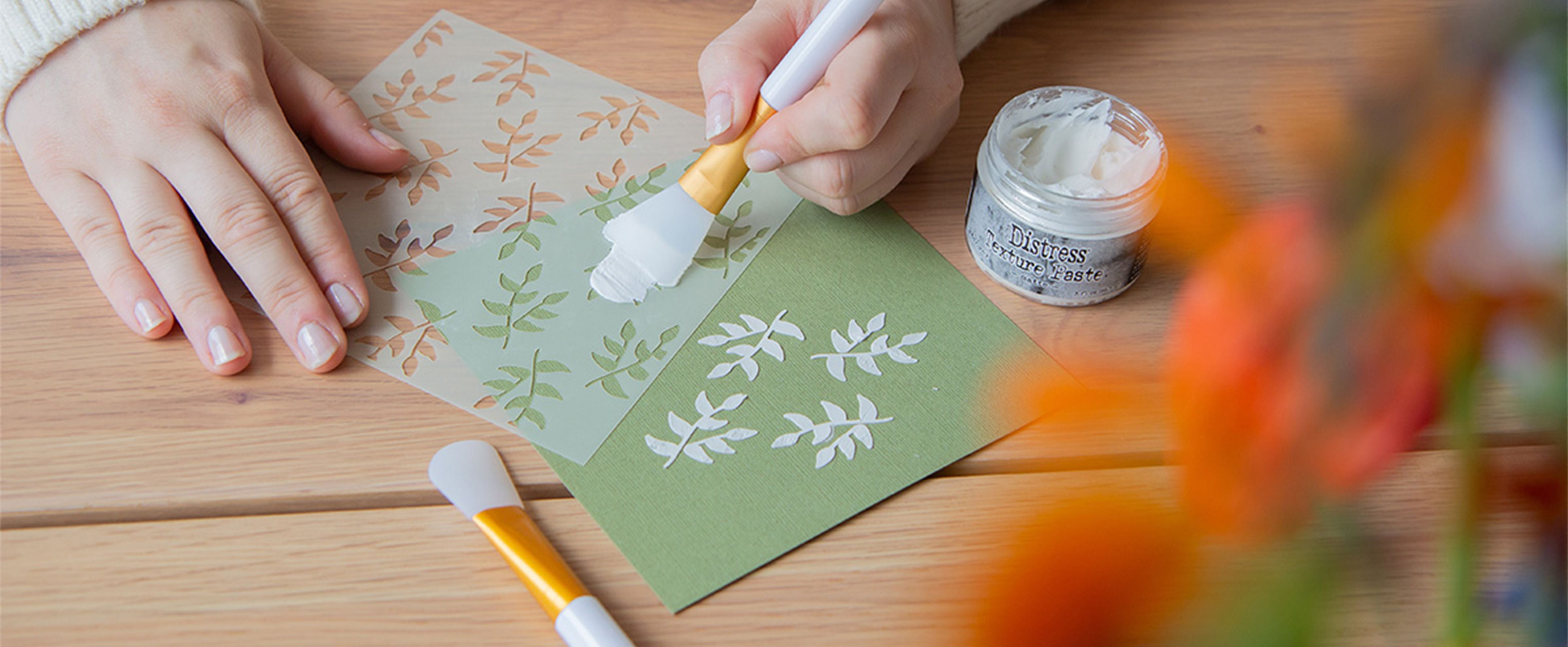 Stamping with stencils and texture paste on green cardstock, while hands apply a leaf pattern for a layered mixed media background.