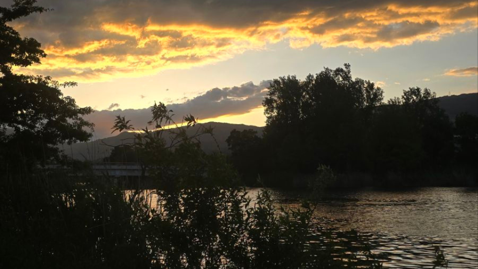 Das goldene Licht der untergehenden Sonne spiegelt sich im fliessenden Wasser. Am Ufer stehen Bäume und Sträucher, während im Hintergrund die Berge der Jurakette zu sehen sind.png