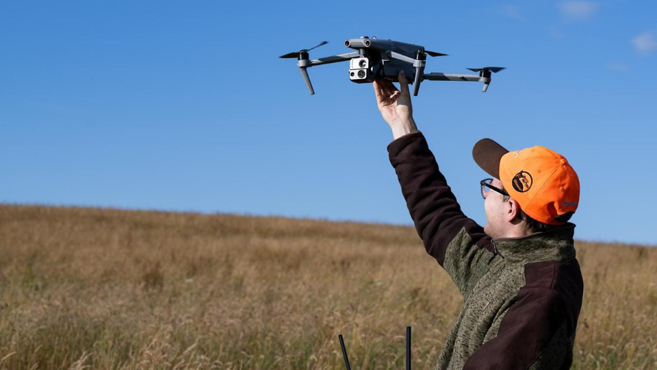 Berufsjäger Gianni Parpan steht mit dem Rücken zur Kamera auf einem Feld und hält eine DJI Matrice 4 Thermal Wärmebilddrohne in den Händen, bereit für die Kitzrettung im Revier.png
