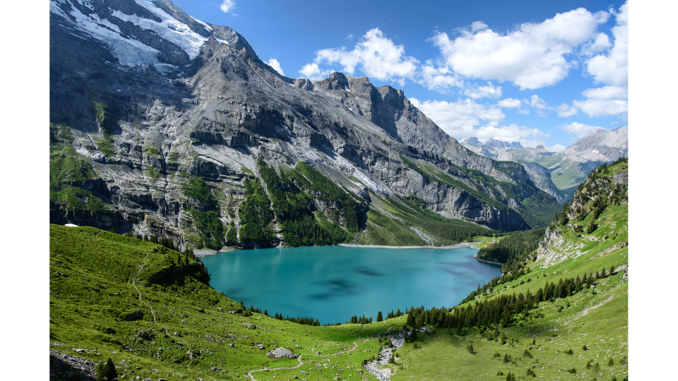 Der türkisfarbene Oeschinensee im Sommer. Das klare Bergwasser ist umgeben von tiefgrünen Wäldern und Bergen mit Schneeresten unter einem leicht bewölkten, blauen Himmel.png