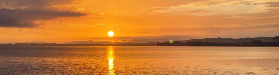 Ein wunderschöner, orangefarbener Sonnenuntergang spiegelt sich im Wasser, mit einer dunklen Landschaft am Horizont beim Welsangeln.png