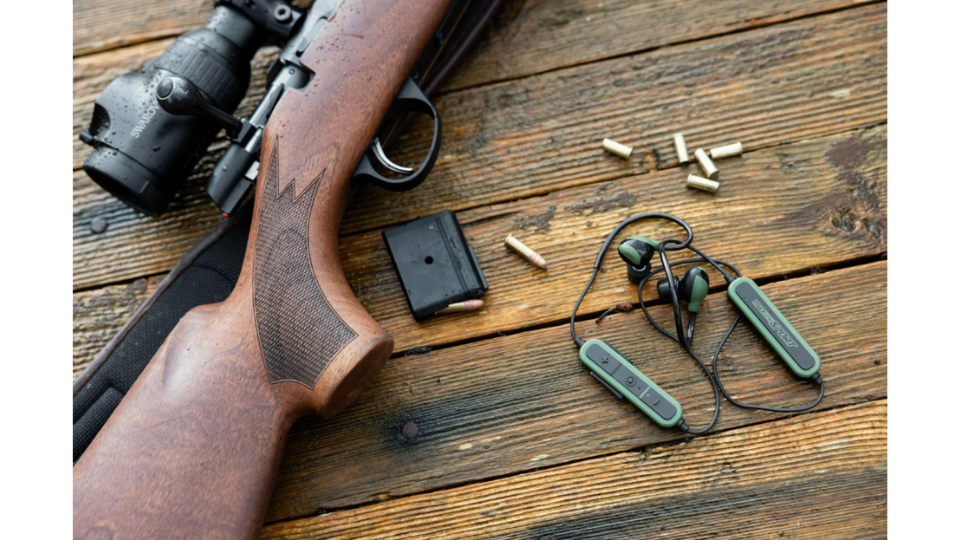 Ein Jagdgewehr mit Zielfernrohr, Munition, ein Magazin und ein aktiver Gehörschutz liegen bereit auf einem Holzboden für das Schiesstraining.png