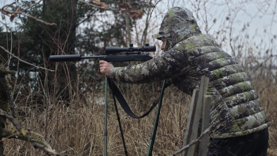 Ein Jäger im Camouflage-Outfit zielt mit seinem auf einem Stativ befestigten Gewehr im Wald, während der Treffsicherheitsnachweis in der Schweiz näher rückt.png
