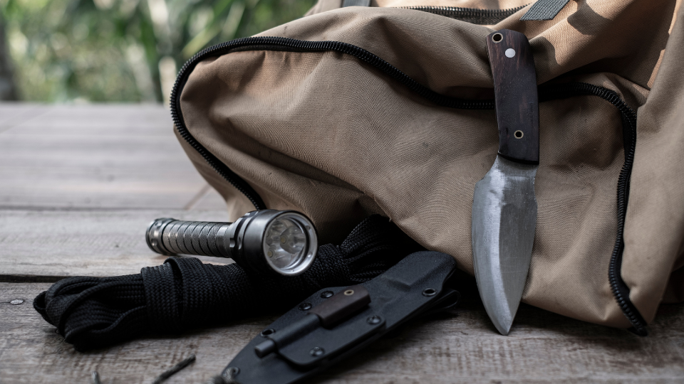 Jagdausrüstung im Check. Ein Jagdmesser mit Holzgriff, eine Taschenlampe und eine Messerscheide liegen bereit vor einem beigefarbenen Jagdrucksack.png