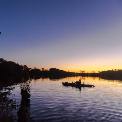 Kajak-Angeln bei Sonnenaufgang. Silhouette eines Anglers in einem Angelkajak auf einem spiegelglatten See am frühen Morgen.png