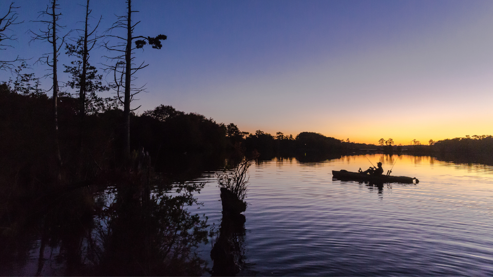 Kajak-Angeln bei Sonnenaufgang. Silhouette eines Anglers in einem Angelkajak auf einem spiegelglatten See am frühen Morgen.png