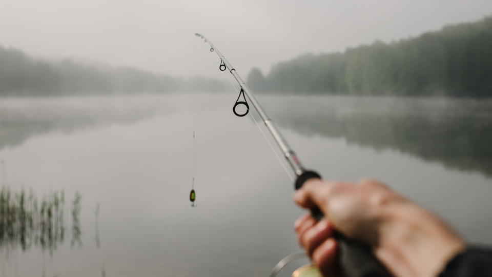 Ultralight-Angeln auf Barsch am frühen Morgen. Ein Angler hält eine feine UL-Rute über einen spiegelglatten, von Nebel umgebenen See im Hochsommer.png