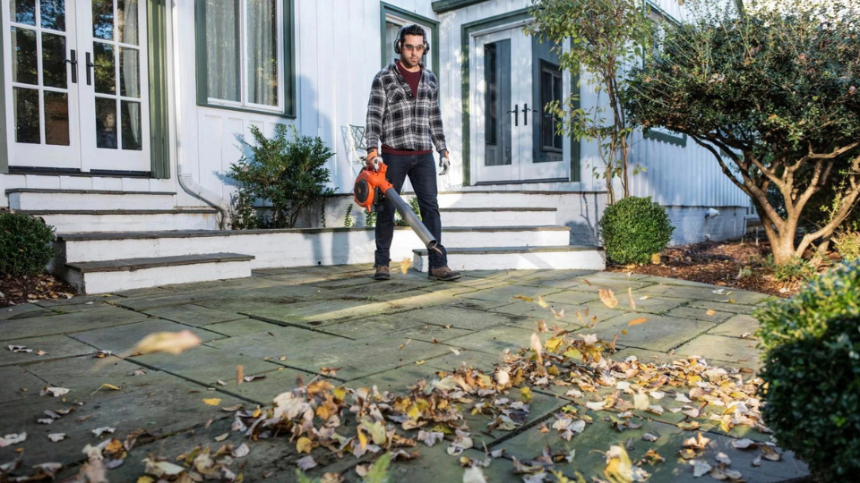 Mann reinigt eine Terrasse vor dem Haus mit einem orangefarbenen Husqvarna Laubbläser im Herbst.png
