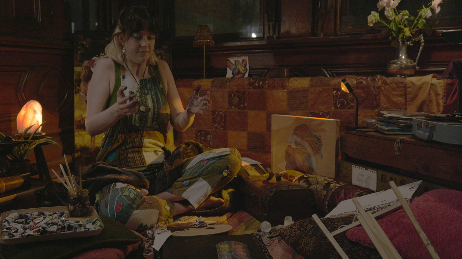 A young woman sits cross-legged on a patterned floor in a dimly lit, bohemian-style room filled with art supplies and decorations. She wears colorful patchwork overalls and holds her paint-covered hands in front of her with a distraught facial expression. An easel is thrown out in front of her, and a wooden box sits beside her crossed legs. Around her are paint tubes, sketchbooks, cushions, and small lamps, while warm lighting and dark wooden walls creating a cozy yet emotionally tense atmosphere.