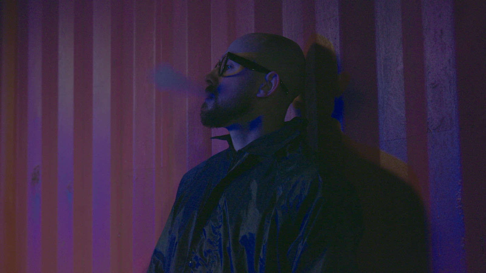 A bald man with glasses and a beard leans against a corrugated metal shipping container under red and blue lighting. He wears a dark jacket and looks upward while exhaling smoke, creating a moody and cinematic atmosphere with strong shadows cast behind him.