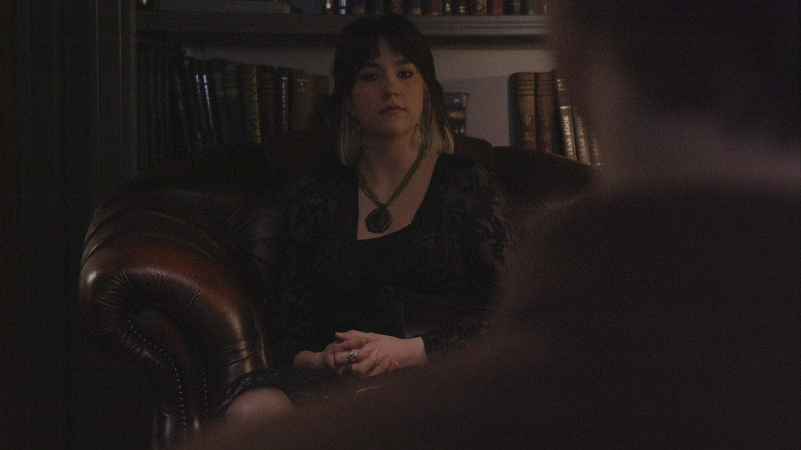 A young woman with dark hair and blonde ends sits in a brown leather armchair in a dimly lit room lined with bookshelves. She wears black clothing and layered jewellery, with her hands folded in her lap as she looks calmly ahead. The blurred silhouette of another person appears in the foreground, creating the feeling of a private conversation or therapy session.