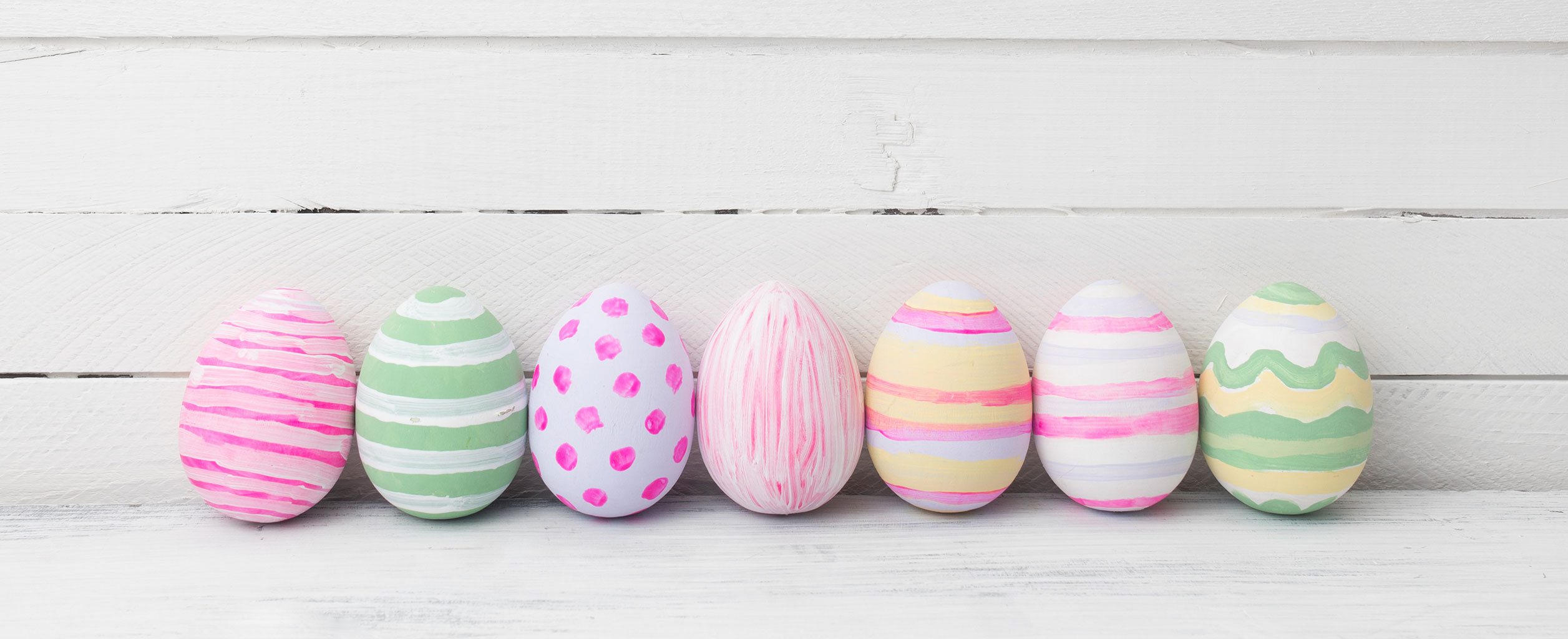 Sieben bemalte Ostereier mit rosa, grünen und gelben Mustern sind vor einem weißen Holzhintergrund aufgereiht.