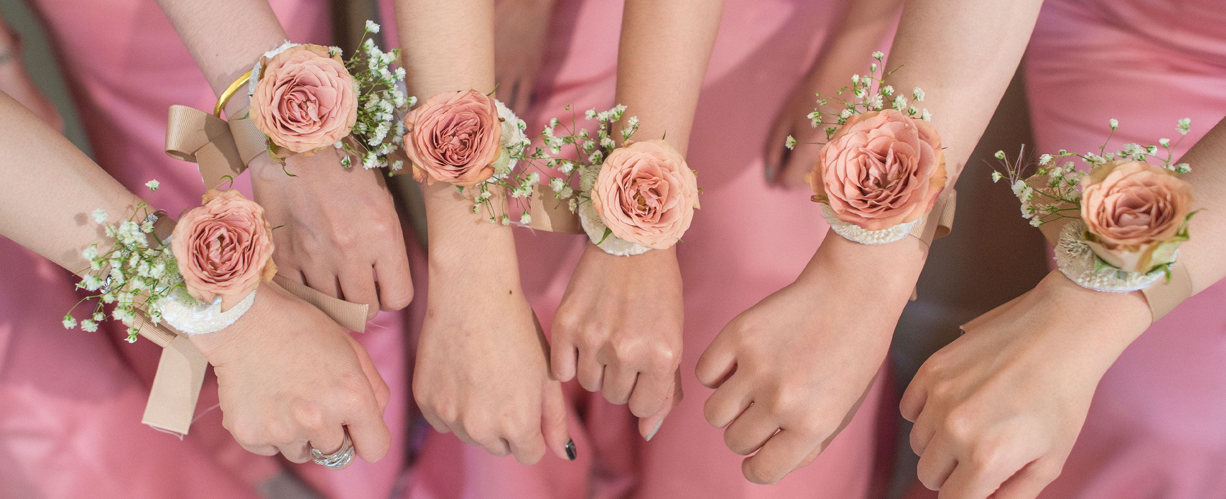 Fünf Personen in rosafarbener Kleidung, die an ihren Handgelenken passende Rosenkorsagen tragen, strecken ihre Hände nach vorne.
