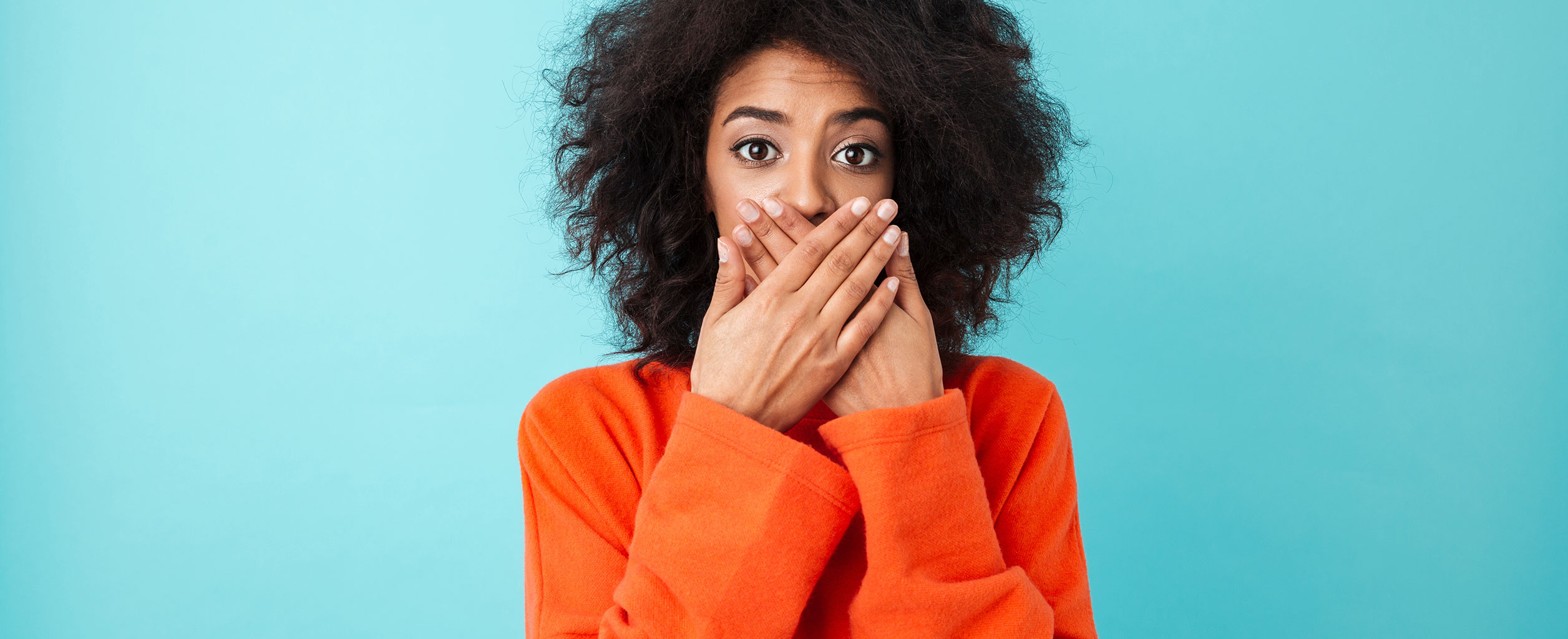 Eine Frau in einem orangefarbenen Pullover, die ihren Mund mit beiden Händen bedeckt, steht vor einem blauen Hintergrund.