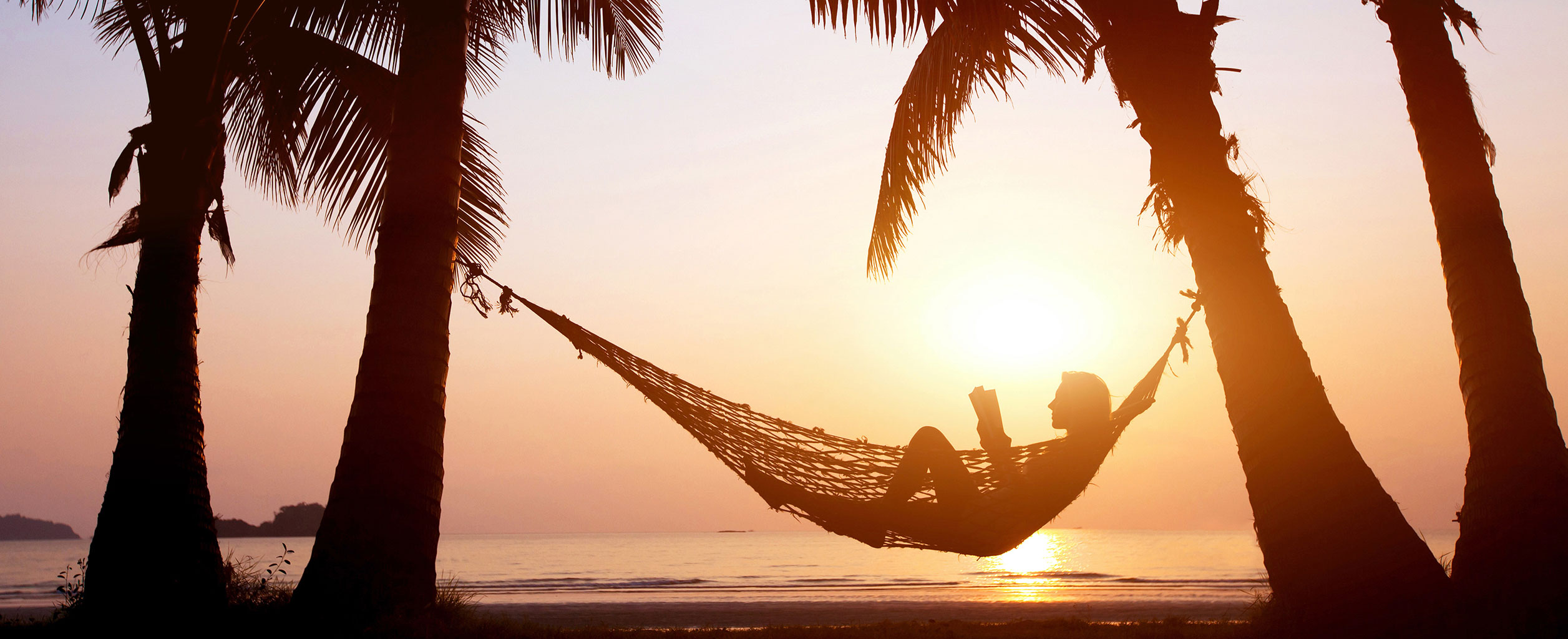 Lesende Person in einer Hängematte zwischen Palmen bei Sonnenuntergang am Strand, mit ruhigem Wasser im Hintergrund.