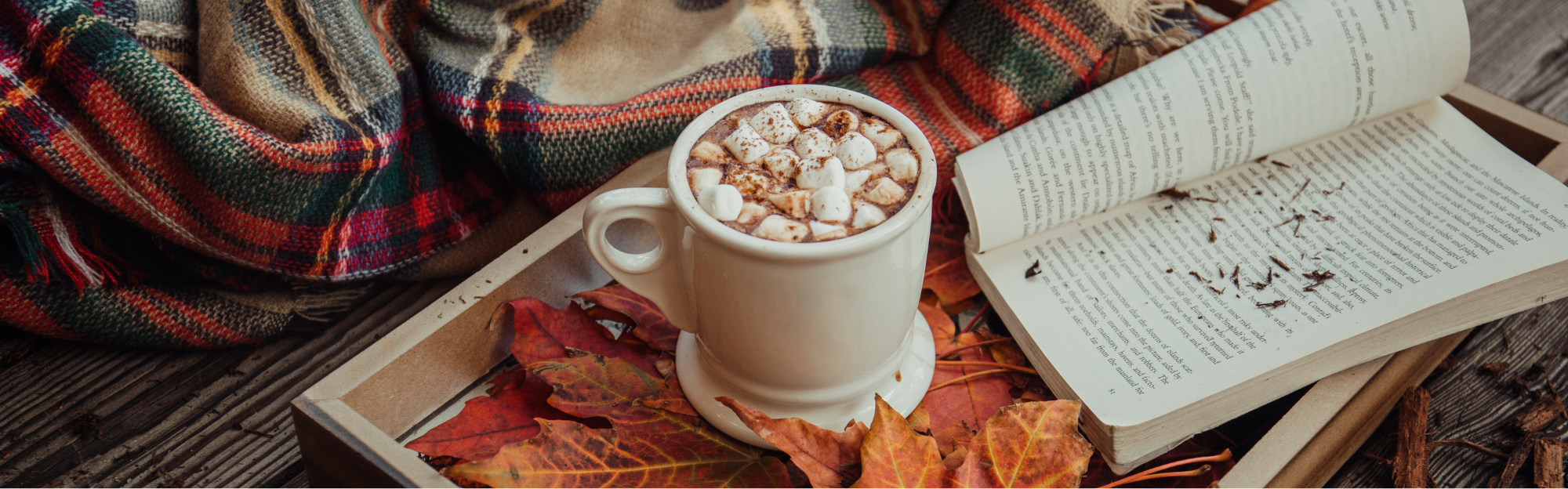 Eine Tasse heiße Schokolade mit Marshmallows steht auf einem Holztablett mit Herbstblättern, neben einem offenen Buch und einer gemütlichen karierten Decke.