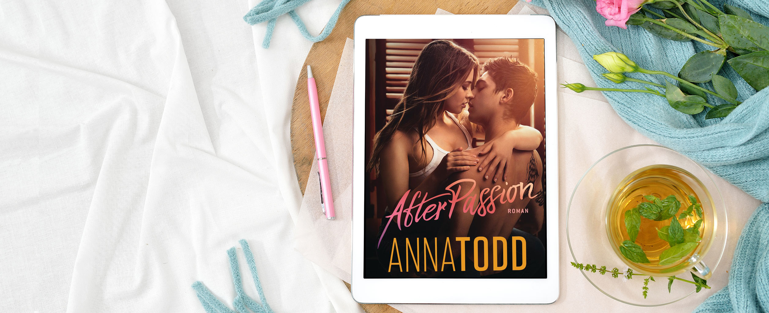Tablet mit dem Buch After Passion von Anna Todd, daneben ein Stift, Tee, eine Rose und ein hellblauer Schal auf einem weißen Tuch.