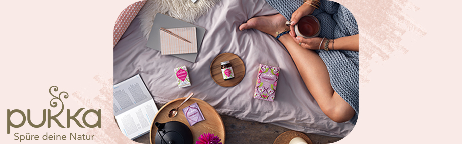 Frau auf einem Bett sitzend mit Tee, Büchern und Pukka-Produkten, von oben gesehen.