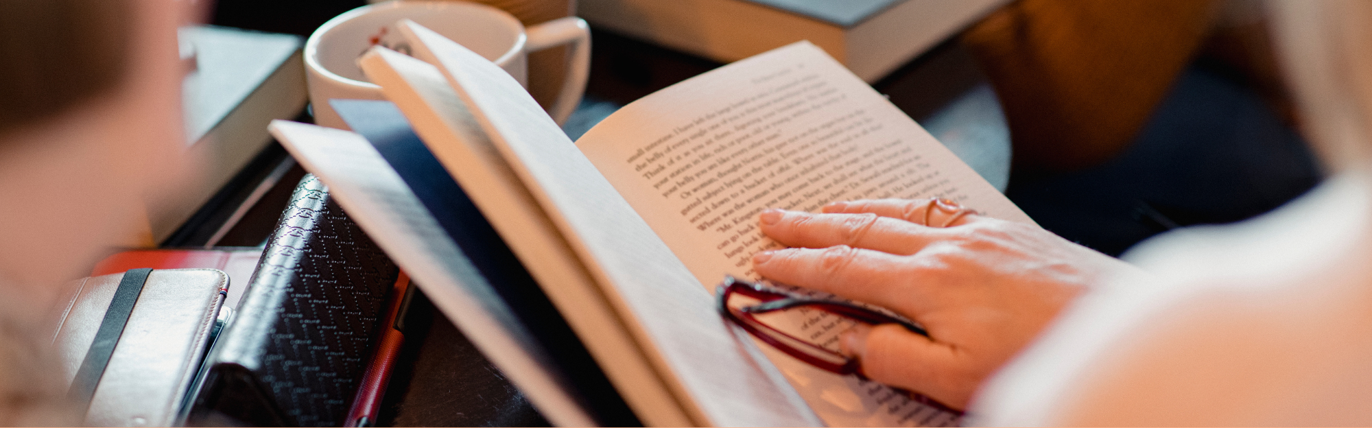 Eine Person hat ein Buch in der Hand und scheint dieses zu lesen. Eine andere Person sitzt daneben, es ist nur eine Silhouette zu erahnen. Es wird der Eindruck eines Buchclubs erweckt. 