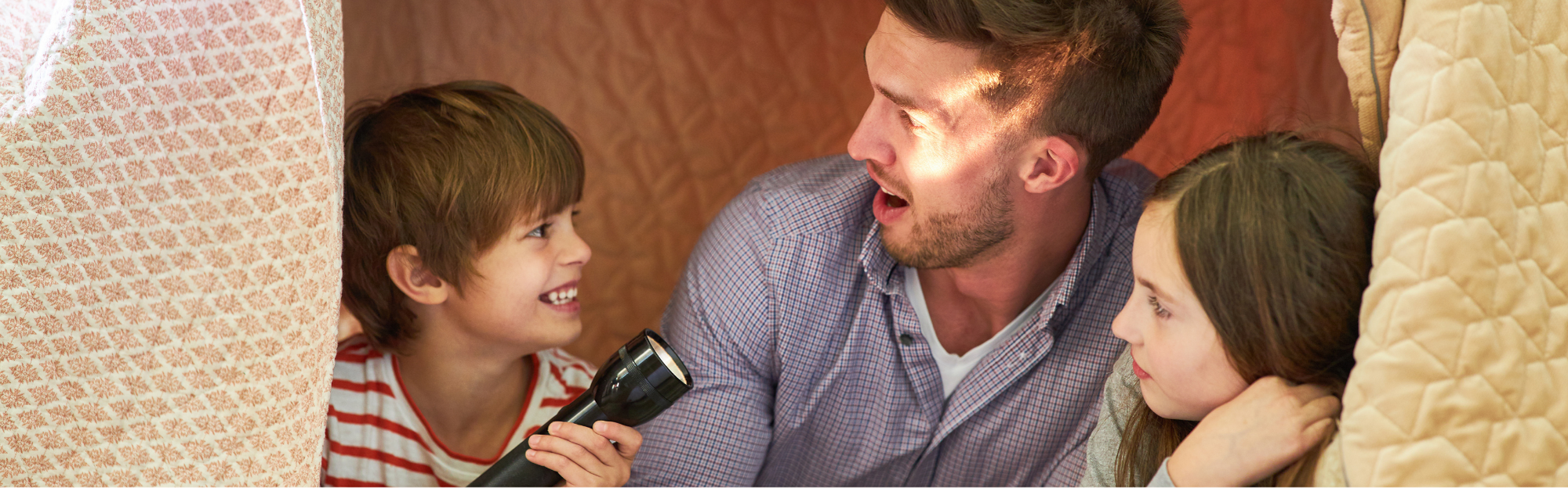 Vater liest zwei Kindern mit Taschenlampe vor, in einem selbst gebauten Zelt.png