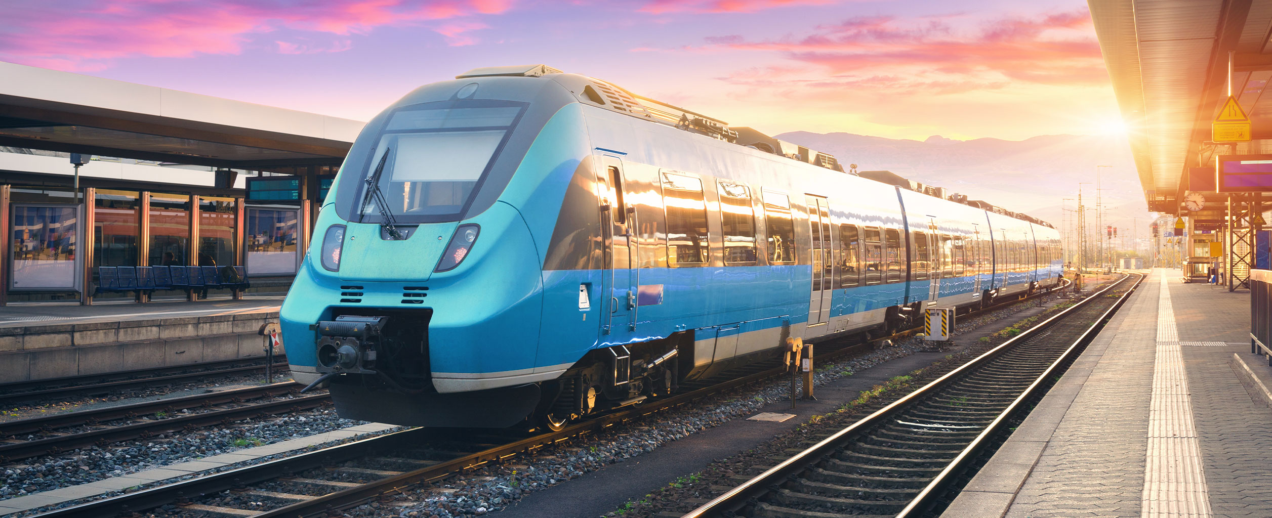 Ein moderner blauer Zug auf einem Bahnsteig bei Sonnenaufgang, mit Gleisen und Bergen im Hintergrund.