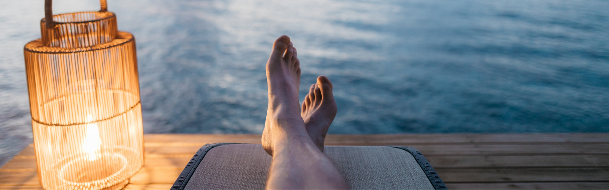 Person auf einem Liegestuhl neben einer beleuchteten Laterne am Wasser liegend, mit ausgestreckten Beinen und Füßen im Bildvordergrund.
