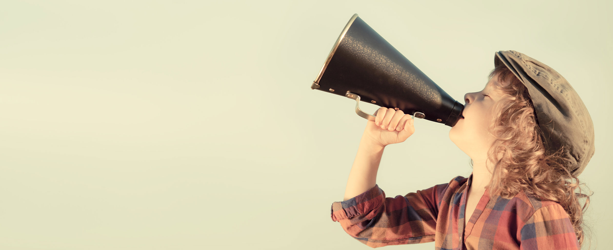 Kind in kariertem Hemd und Mütze, das ein schwarzes Megaphon hält und vor einem schlichten Hintergrund spricht.