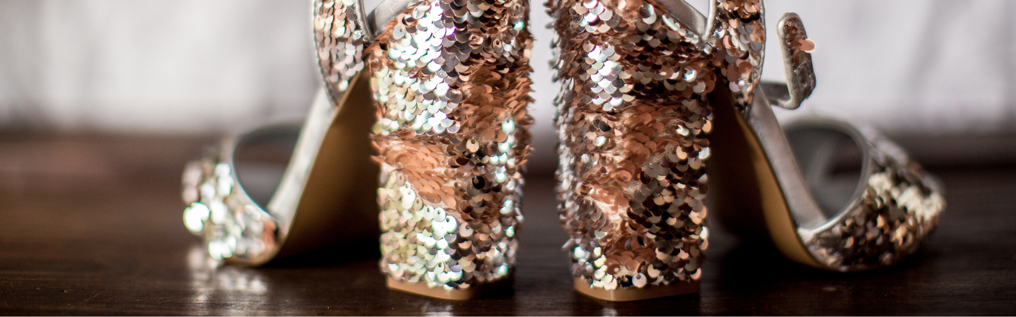 Nahaufnahme eines Paars hochhackiger Schuhe mit glänzenden, roségoldenen Pailletten auf einem Holzboden.