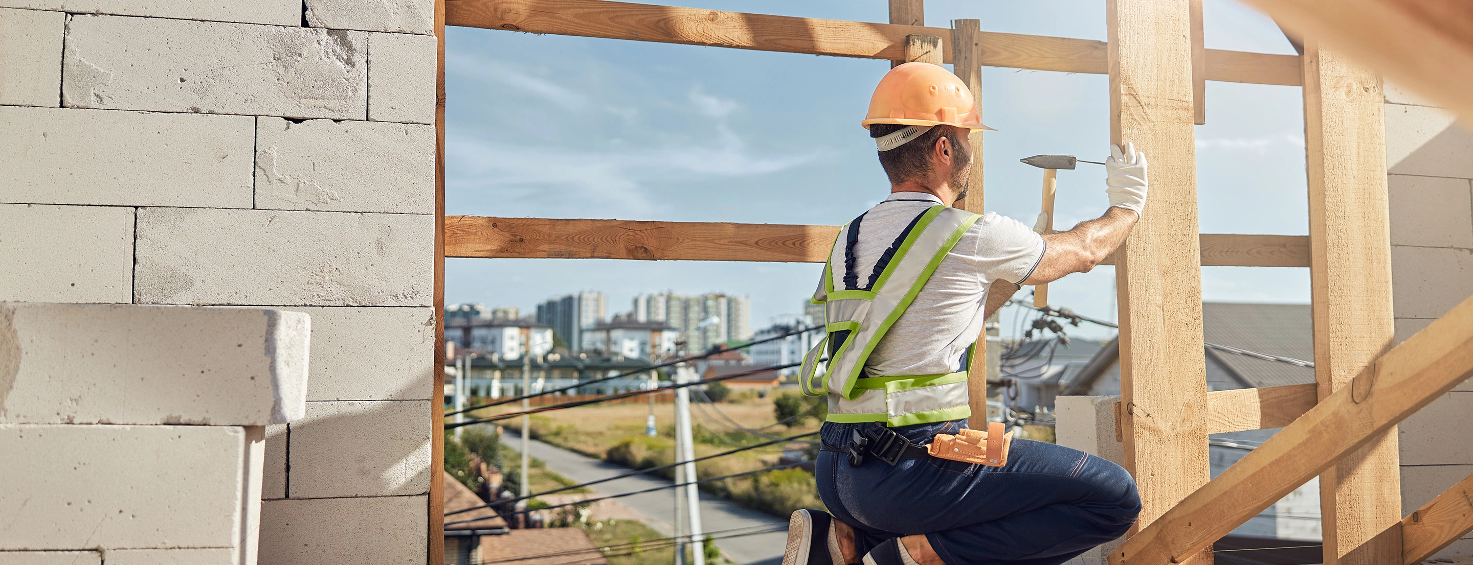 Ein arbeitender Mann auf einer Baustelle, haut einen Nagel in einen Balken. 