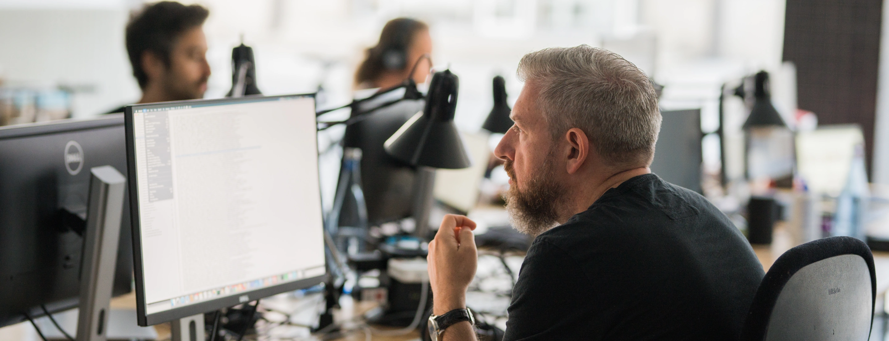 Man sitting at a desk in front of a monitor, with other people working in the office in the background