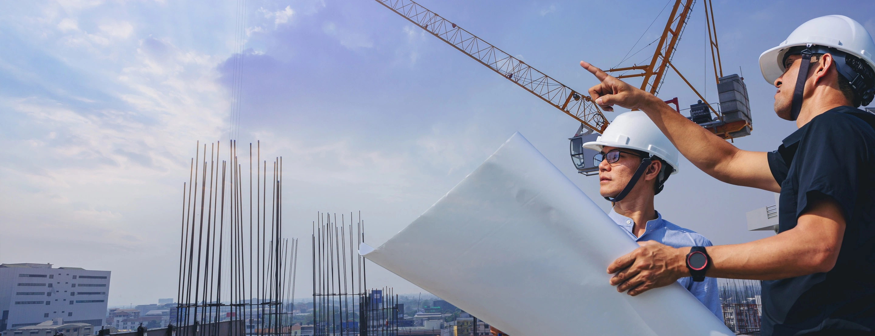 2 Menschen auf einer Baustelle besprechen etwas, im Hintergrund ist die Baustelle und ein Kran zu sehen.