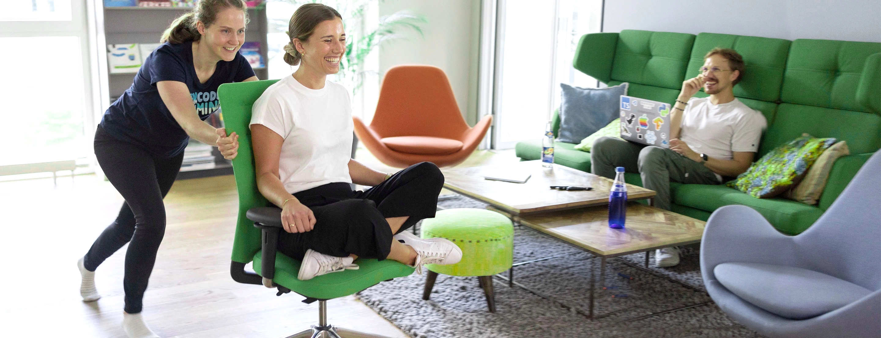 A person laughing being rolled around on a chair by another person, with yet another person laughing in the background.