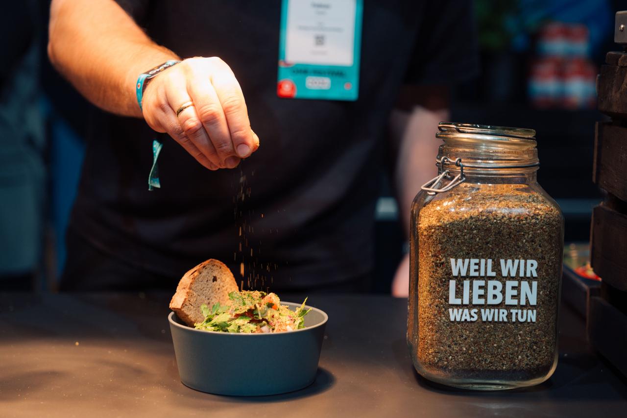 Ein Salat mit Brot in einem Mehrwegbehälter. Im Hintergrund ist eine Person zu sehen, die Gewürze über die Speise mit dem Finger verteilt.