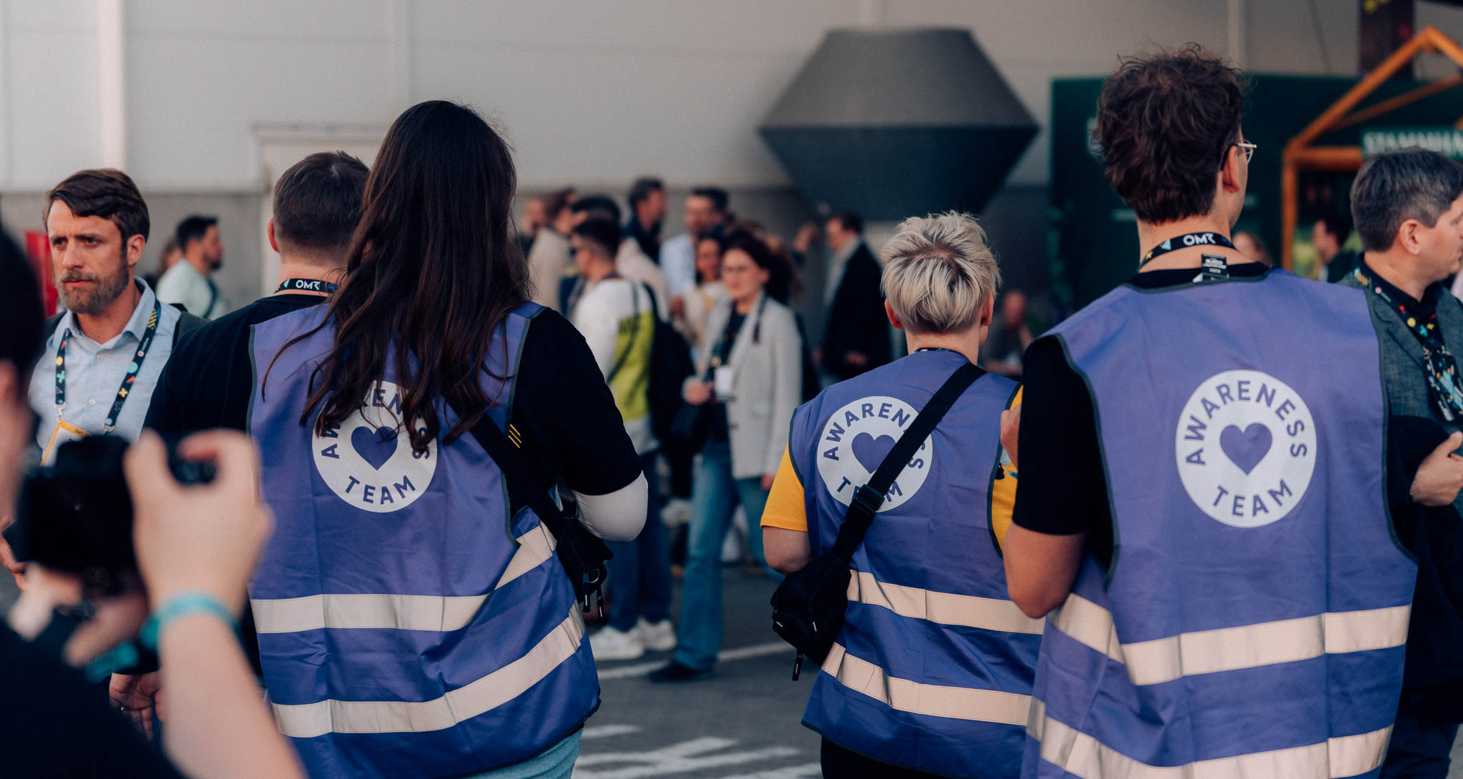 Blick von hinten auf drei Menschen, die alle eine lila Weste anhaben mit der Aufschrift "Awareness Team".