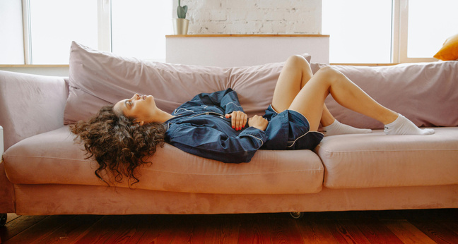 Eine junge Frau liegt in bequemer Kleidung zusammengerollt auf einem rosa Sofa. Sie hält sich den Bauch, wirkt erschöpft und ruht sich aus. Tageslicht fällt durch große Fenster in den hellen Raum. Die Szene vermittelt Rückzug, Schmerzen und das Bedürfnis nach Ruhe – typisch für Menstruationsbeschwerden.