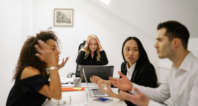 Vier Personen in einem Büro diskutieren heftig, während eine Frau im Hintergrund mit geschlossenen Augen und Händen an den Schläfen sichtlich gestresst wirkt.