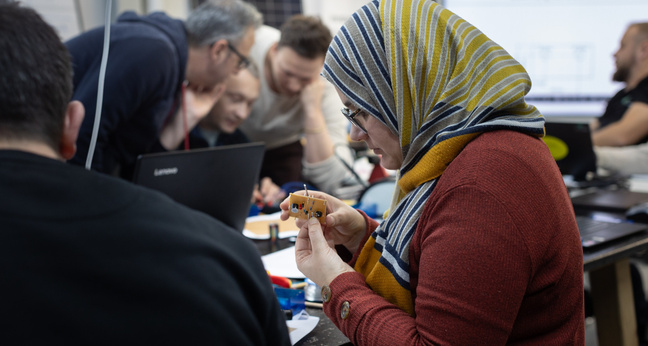 In einem Schulungsraum sitzt eine Frau mit gestreiftem Kopftuch und konzentriertem Blick an einem Tisch und hält ein elektronisches Bauteil in der Hand. Um sie herum arbeiten weitere Teilnehmende mit Laptops und Werkzeugen – eine Szene aus einem praktischen Ausbildungskurs zur Elektrofachkraft für das Klimahandwerk.