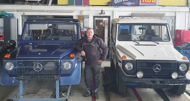 In einer Kfz-Werkstatt steht Patrick Lippick in schwarzer Arbeitskleidung zwischen zwei Mercedes G-Klassen. Die Fahrzeuge sind teils restauriert und in Blau und Beige lackiert. Die Aufnahme zeigt eine typische Werkstattumgebung mit Werkzeug, Reifenstapel und Hebebühne – ein authentischer Einblick in den Alltag im Azubi-Recruiting im Handwerk.