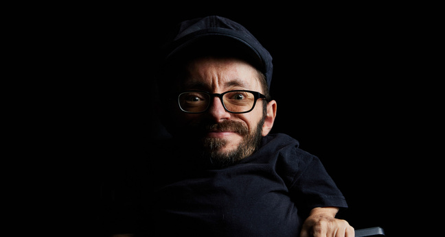 Ein Porträt von Raúl Krauthausen vor schwarzem Hintergrund. Er trägt Brille, Mütze und ein dunkles Oberteil, lächelt leicht und schaut direkt in die Kamera. Die Aufnahme ist ausdrucksstark und reduziert – der Fokus liegt ganz auf seiner Person.