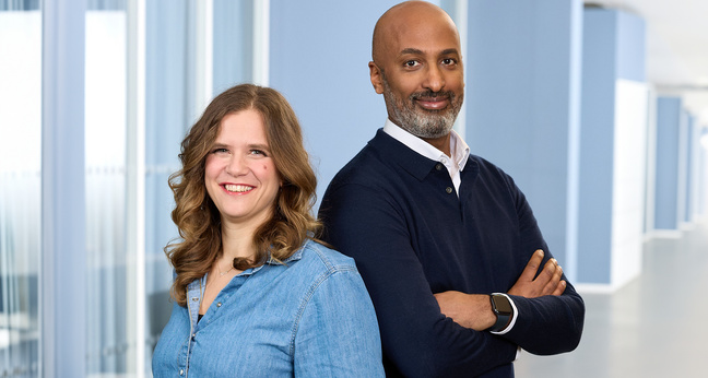 Ein Mann im dunklen Pullover mit verschränkten Armen steht neben einer Frau in Jeanshemd, beide lächeln in die Kamera. Der helle Büroflur im Hintergrund unterstreicht die professionelle Atmosphäre – Symbol für moderne Jobsharing-Modelle in Unternehmen.